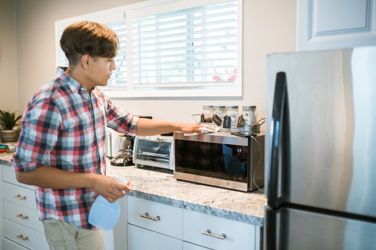 A man is cleaning a microwave in his modern kitchen using a spray bottle and cloth.